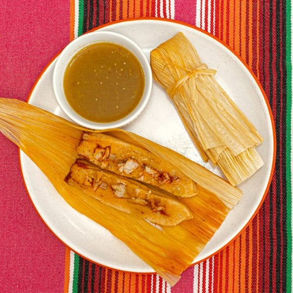La Patrona's tamales, one unwrapped with pork and two wrapped, served with green salsa on a colorful Mexican placemat.