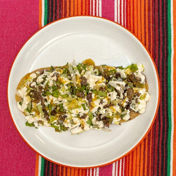 La Patrona's huarache topped with meat, cheese, lettuce, and sauce on a colorful striped background.