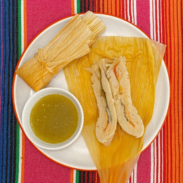 La Patrona tamales, one unwrapped, with green salsa on a white plate over a colorful striped tablecloth.