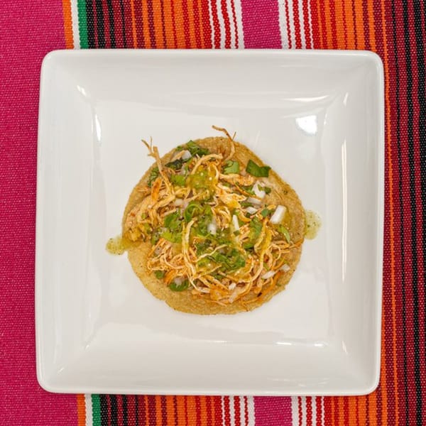 A La Patrona chicken tostada with cilantro and onions on a white plate over a colorful striped tablecloth.