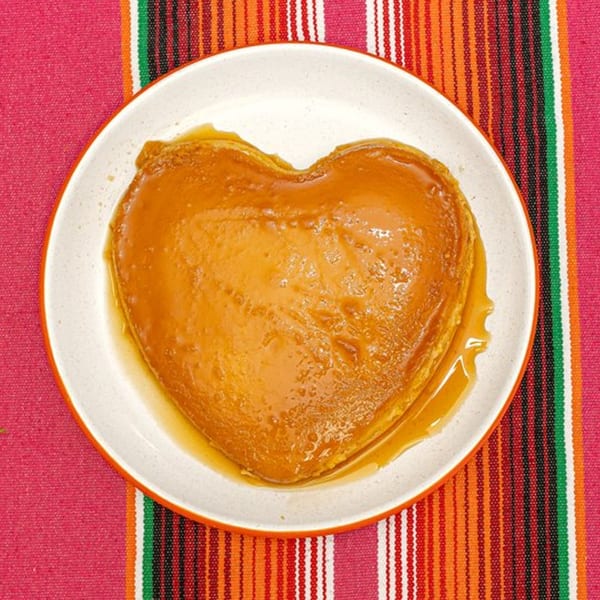 Heart-shaped flan with caramel sauce on a white plate, atop a colorful striped Mexican serape.