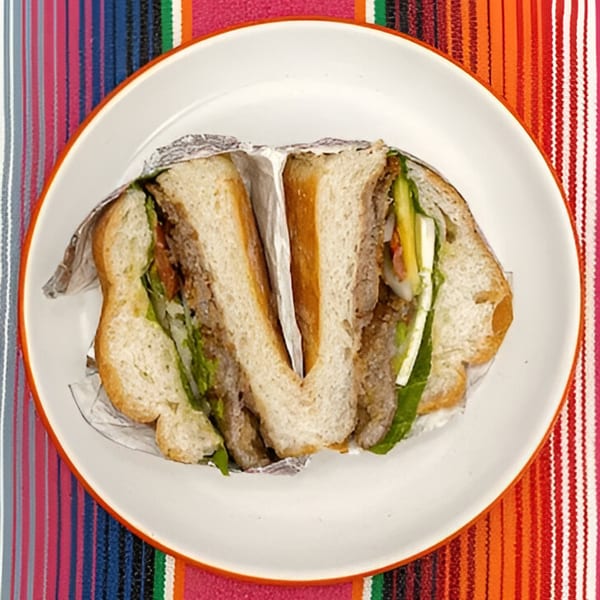 A Mexican torta sandwich, cut in half and wrapped in foil, on a white plate over a colorful striped tablecloth.
