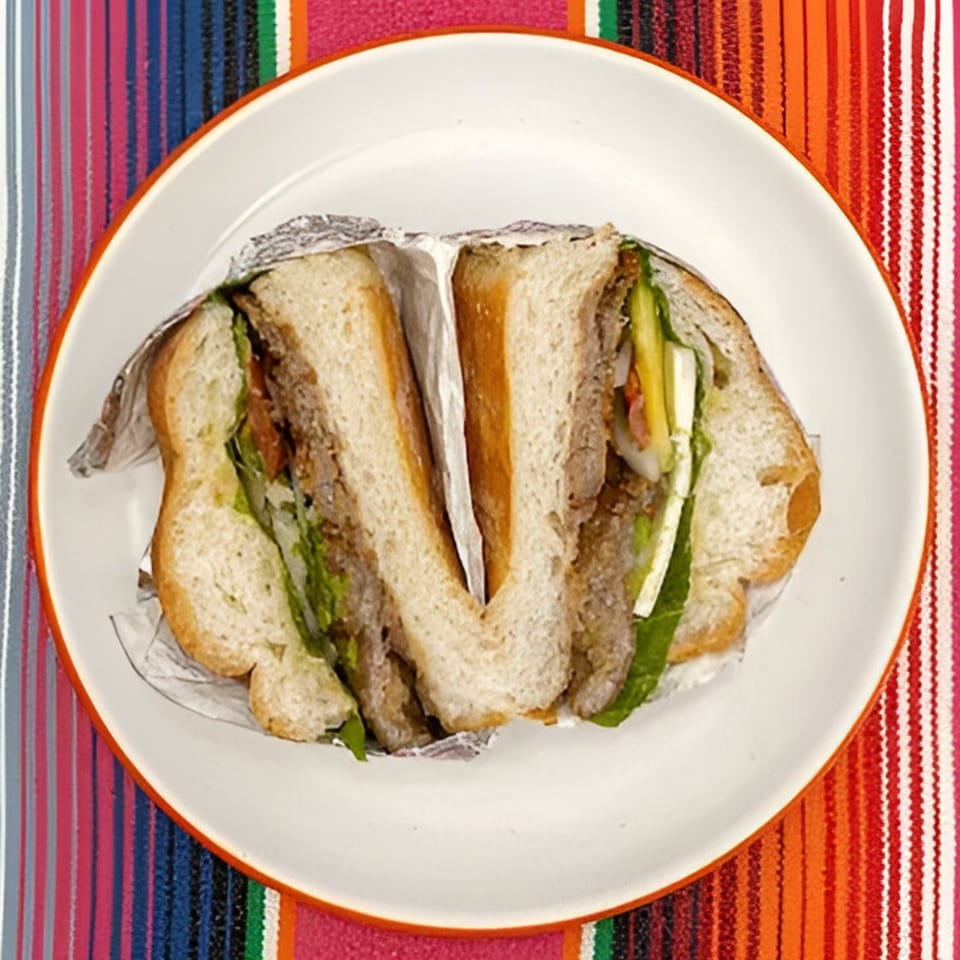 A Mexican torta sandwich, cut in half and wrapped in foil, on a white plate over a colorful striped tablecloth.