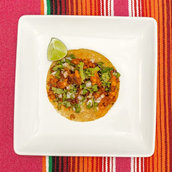 A single La Patrona Mexican taco with meat, onions, cilantro, and a lime wedge on a white plate.