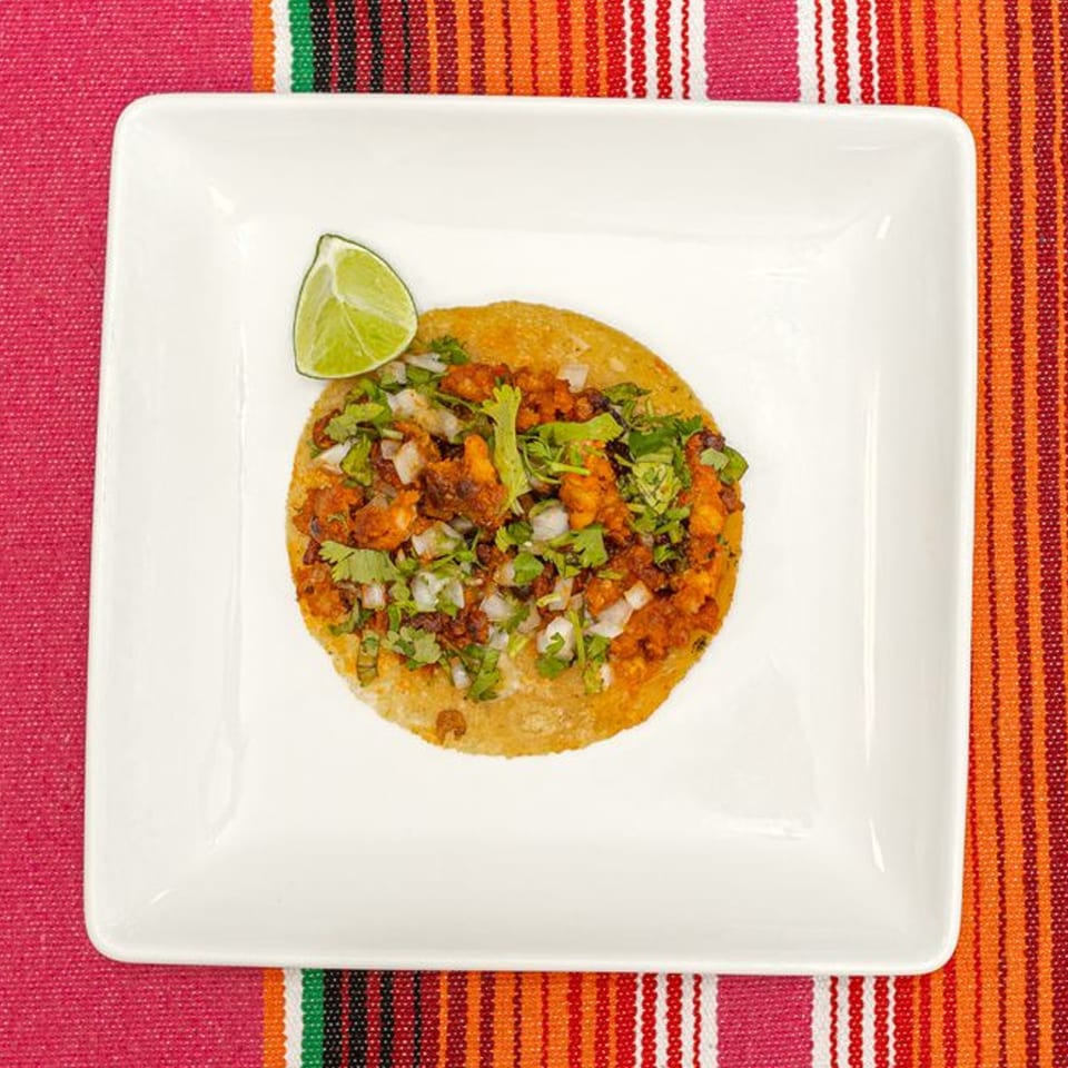 A single La Patrona Mexican taco with meat, onions, cilantro, and a lime wedge on a white plate.