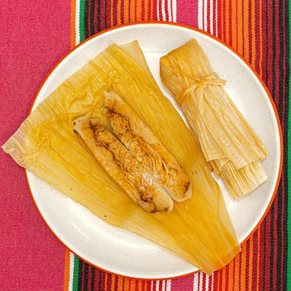 Two La Patrona tamales, one unwrapped revealing chicken filling, on a white plate with a colorful striped tablecloth.